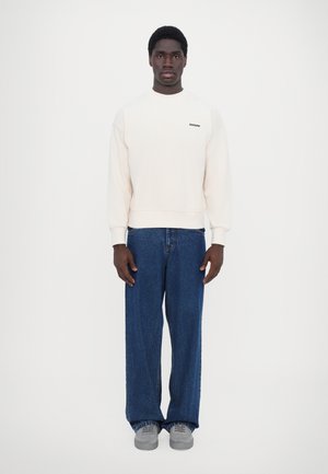 Sweatshirt creme com um decote redondo e punhos canelados, combinado com jeans azuis de corte solto e ténis cinzentos, de pé contra um fundo branco.