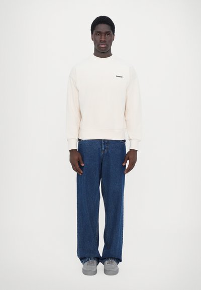 Sweatshirt crème à col rond et manches côtelées, associé à un jean bleu ample et des baskets grises, se tenant devant un fond blanc.