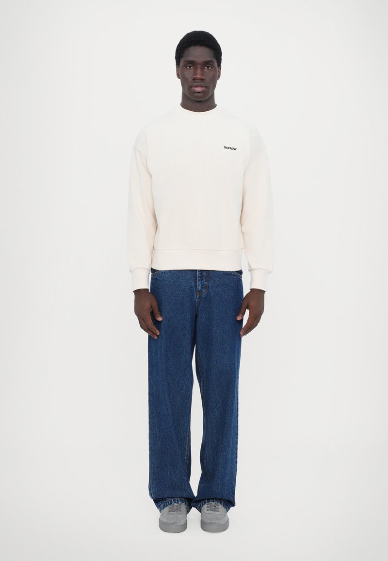 Sweatshirt crème à col rond et manches côtelées, associé à un jean bleu ample et des baskets grises, se tenant devant un fond blanc.