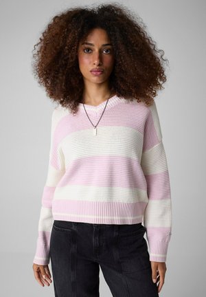 Donna con capelli ricci voluminosi indossa un maglione a righe rosa e bianco e pantaloni neri, in piedi davanti a uno sfondo grigio uniforme.