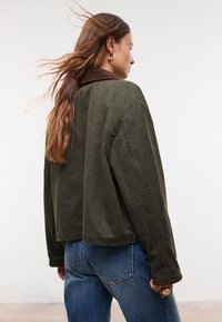 Donna con lunghi capelli castani che indossa orecchini a cerchio dorati, una giacca verde scuro con colletto marrone e jeans blu, che sta in piedi su uno sfondo bianco.