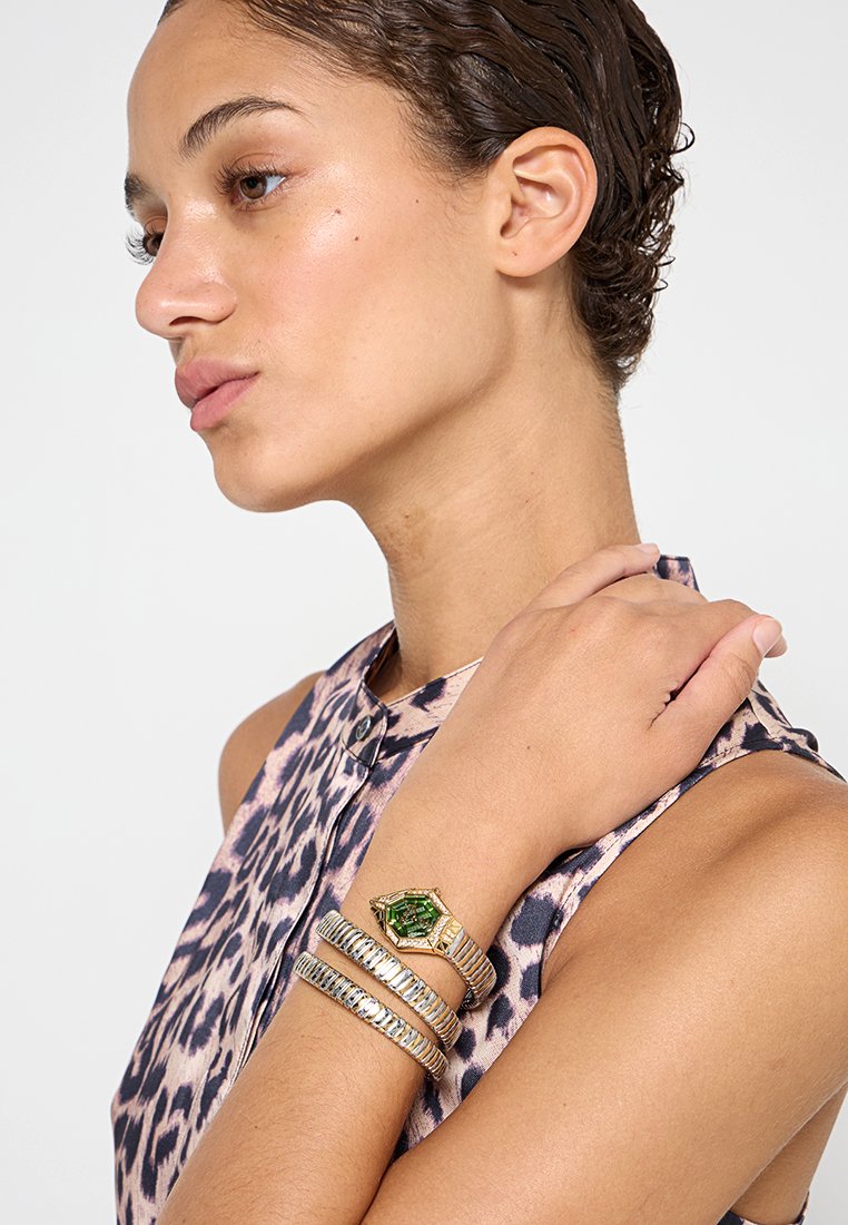 Pulsera de piedras preciosas hexagonales en oro y verde con pulseras de plata y oro. Presentan superficies texturizadas y lisas. Llevada en una muñeca descubierta.