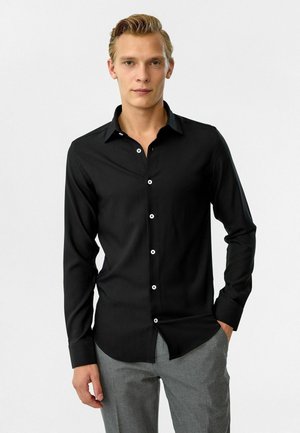 Zwarte button-up shirt met een kraag, lange mouwen en een gladde textuur. Het heeft witte knopen en een getailleerde pasvorm, gecombineerd met grijze broeken.