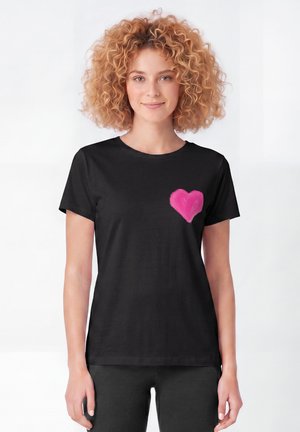 Vrouw met krullend haar die een zwart T-shirt draagt met een felroze hart op de linkerborst, staand tegen een effen witte achtergrond.