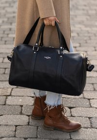 Zwarte leren reistas met twee bovenhandvatten, zilverkleurige hardware, subtiel logo aan de voorkant en een gestructureerd oppervlak. Draagt bij een denim outfit en bruine laarzen.