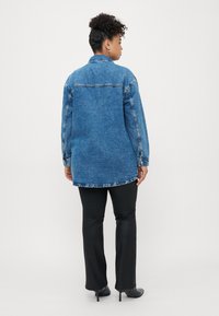 Woman with curly hair wearing blue oversized denim jacket, black fitted pants, and black pointed heels, shown from the back standing on a white background.