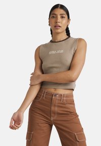 Brązowy prążkowany crop top z kontrastowym ciemnym dekoltem, z bia�łym logo tekstowym. W połączeniu z brązowymi spodniami typu high-waisted z kieszeniami cargo.