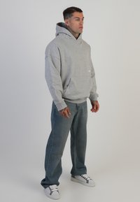 Felpa grigia in cotone con tasca frontale, caratterizzata da una vestibilità comoda e un ampio cappuccio. Indossata con jeans azzurri chiari e sneakers bianche.