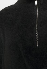 Sudadera de felpa negra con cuello alto, que presenta una cremallera metálica y una tela suave y texturizada. No se observan patrones ni acentos adicionales.