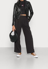 Nainen, jolla on päällään musta nahkatakki, musta crop-top, mustat leveälahkeiset housut, ruskeat lenkkarit ja joka pitää kädessään mustaa tikattua käsilaukkua.