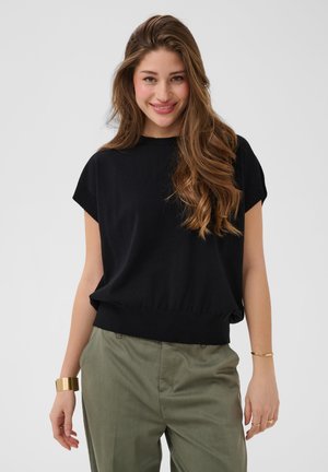 Jonge vrouw met lang golvend bruin haar, een zwart topje met korte mouwen, olijfgroene broek en gouden armbanden glimlachend tegen een effen achtergrond.
