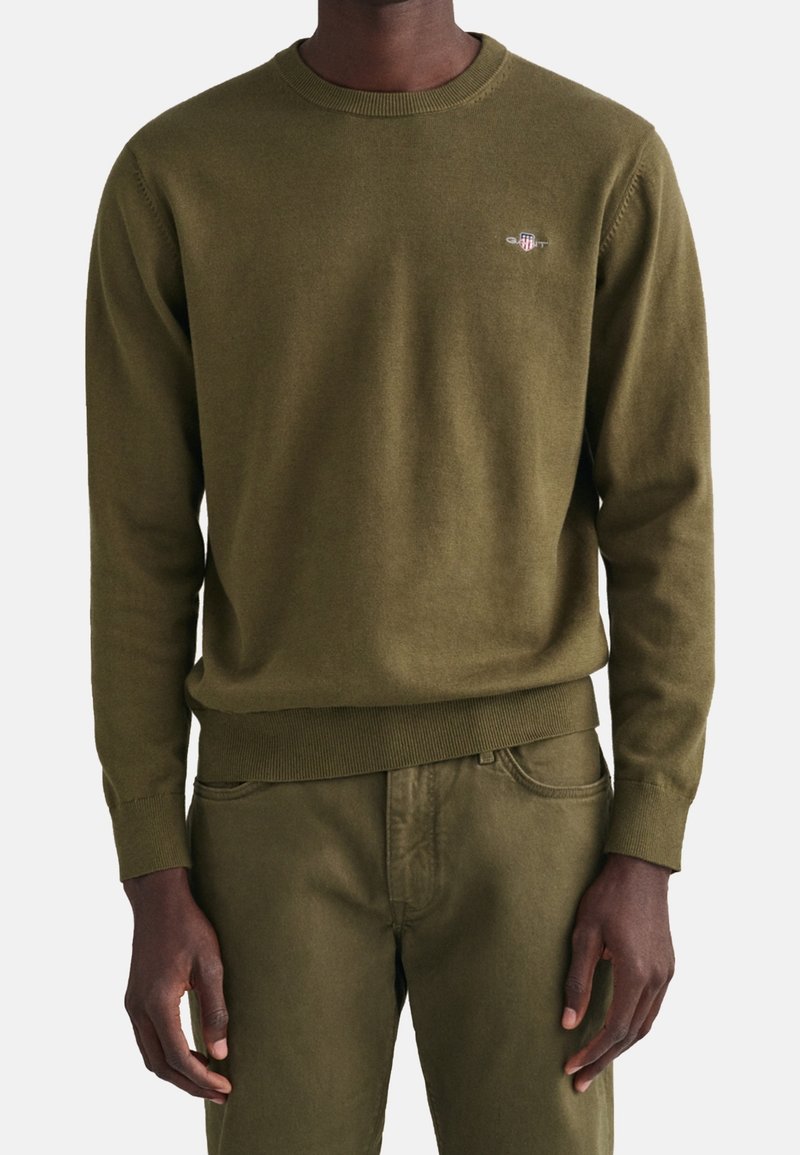 Olivgrüner gestrickter Pullover mit Rundhalsausschnitt und gerippten Bündchen. Subtiles Logo auf der Brust und eine weiche Textur, kombiniert mit einer passenden grünen Hose.