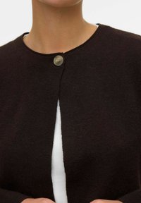 Brauner Strickcardigan mit rundem Ausschnitt, der mit einem einzelnen Knopf schließt und eine strukturierte Oberfläche hat. Über einem weißen Oberteil getragen.
