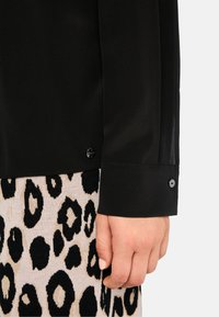 Schwarze Bluse mit langen Ärmeln und einem Bündchen. Verfügt über ein glänzendes Knopfdetail. Getragen über einem Strickrock mit Leopardenmuster in Schwarz und Beige.
