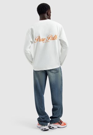 Mann trägt ein weißes Langarmshirt mit dem Schriftzug „Pure Path“, blaue Jeans und weiße Sneaker mit orangefarbenen und schwarzen Akzenten, steht mit dem Rücken zur Kamera.