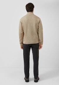 Ein Mann steht mit dem Rücken zur Kamera, trägt einen beigen Pullover, dunkle graue Hosen und braune Schuhe vor einem einfarbigen hellen Hintergrund.