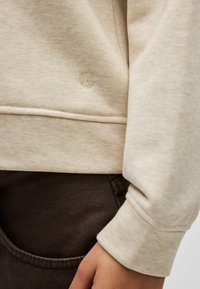 Φωτεινή μπεζ φούτερ κατασκευασμένη από μαλακό ύφασμα, με μικρό κεντητό λογότυπο, ορατή ραφή και ριμπ μανσέτες, συνδυασμένη με καφέ παντελόνι.