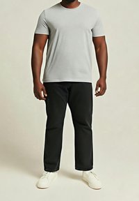 Homme portant un t-shirt à manches courtes gris clair, un pantalon noir et des baskets blanches, debout devant un fond clair uni.