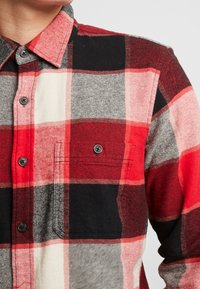 Gros plan sur une personne portant une chemise à carreaux rouge, noire, blanche et grise avec une poche poitrine et un col.