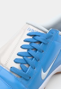 Blau-weißer Sportschuh mit strukturiertem synthetischem Obermaterial, flachen blauen Schnürsenkeln und einem weißen Nike-Swoosh. Die Details umfassen eine verstärkte Zehenkappe.