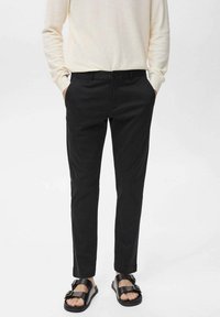 Pantaloni neri slim-fit con tasche frontali, realizzati in tessuto liscio. Abbinati a sandali scuri e a un top a maniche lunghe di colore chiaro.
