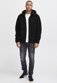 Black, fluffy fleece hoodie with a front zipper, adjustable drawstring hood, and two front pockets, paired with slim grey jeans and black sneakers.