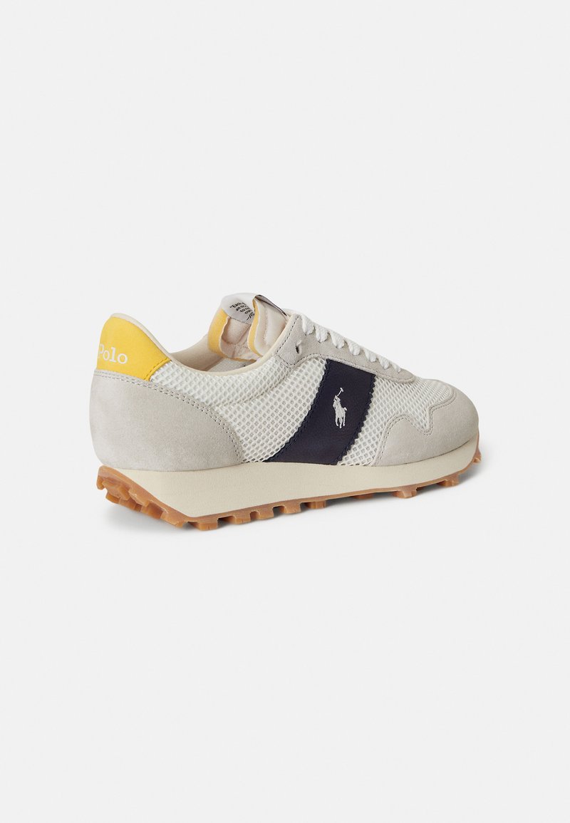 Baskets gris et beige avec panneaux en mesh, accents en suède et logo bleu foncé. Détail jaune au talon et semelle en caoutchouc texturée avec adhérence.