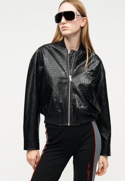 Γυναίκα που φοράει υπερμεγέθη μαύρο δερμάτινο μπουφάν με σχέδιο κροκόδειλου, μεγάλα μαύρα γυαλιά ηλίου και μαύρο παντελόνι με γκρι και κόκκινες ρίγες στα πλάγια.