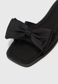 Sandalie negre cu un accent mare din satin sub formă de fundă, vârf pătrat, textură netedă și talpă interioară căptușită, având o talpă de cauciuc plată.