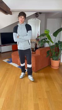 Sweatshirt de zip cinza claro com detalhes pretos, combinado com calças pretas com riscas brancas. Ténis prateados sobre um chão de madeira.