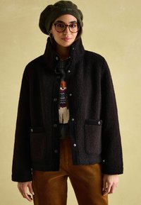 Czarna polarowa kurtka z wysokim kołnierzem, zatrzaskami i kieszeniami naszytymi. Nosi się ją na ciemny wzorzysty sweter i zestawia z spodniami z sztruksu.