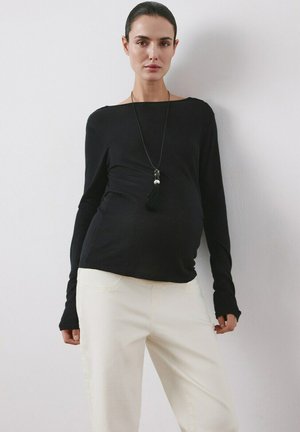 Kobieta w ciąży ubrana w czarną bluzkę z długim rękawem, białe spodnie i długi czarny naszyjnik, stojąca na tle jasnej, gładkiej ściany.