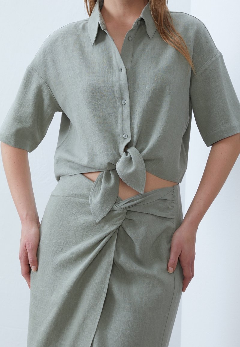 Femme portant une chemise boutonnée à manches courtes vert clair nouée à la taille et une jupe assortie avec un détail de nœud froncé à l'avant.