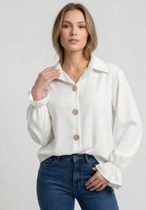 Frau trägt eine weiße, geknöpfte Bluse mit Rüschensäumen an den Manschetten und blaue Jeans, posiert mit einer Hand am Kragen und der anderen in der Tasche.