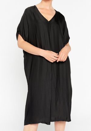 Frau trägt locker sitzendes, kurzärmeliges schwarzes Kleid mit V-Ausschnitt, steht mit verschränkten Händen an der Taille vor weißem Hintergrund.