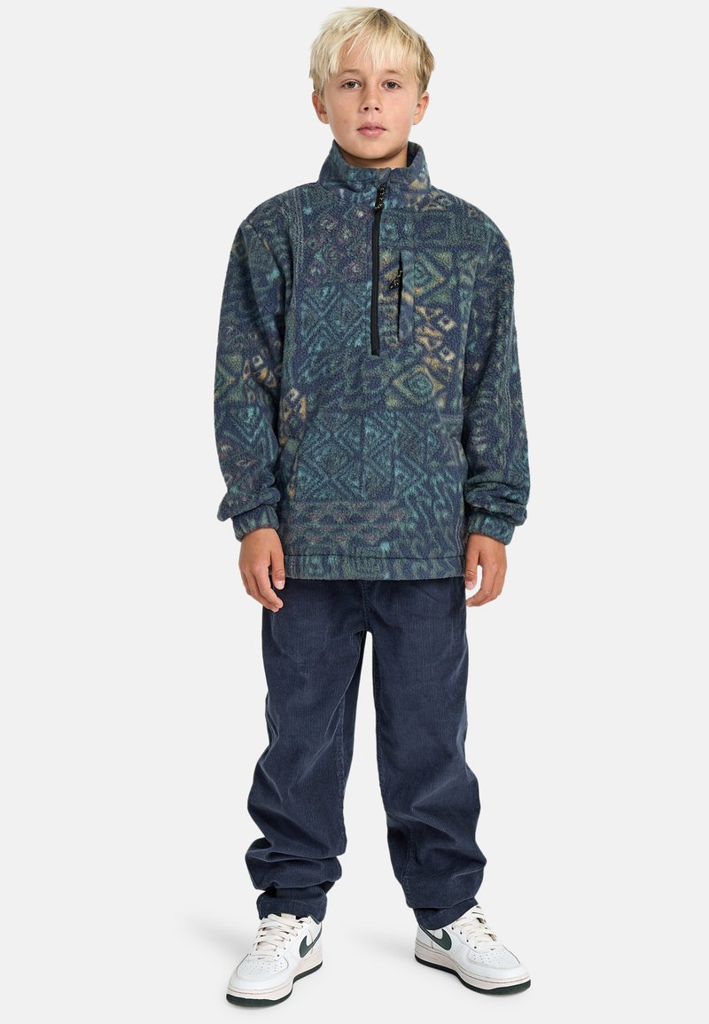 Fleece-Pullover in blauen und grünen geometrischen Mustern, mit einem Halbzipp-Frontreißverschluss, großer Fronttasche und gerippten Bündchen, kombiniert mit navyfarbenen Hosen.