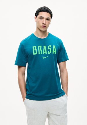 Giovane uomo con capelli corti e ricci che indossa una maglietta turchese "BRASA" e pantaloni grigio chiaro, in piedi con le mani nelle tasche su sfondo bianco.