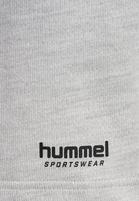 Witte katoenen stof met een ribbelstructuur, met zwarte bedrukte tekst "hummel" en "SPORTSWEAR" aan de onderrand.