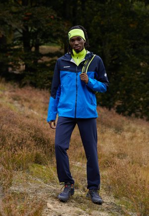 Blue and black waterproof jacket with a high collar, paired with a yellow neck gaiter, navy pants, and gray hiking shoes.