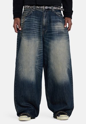 Persona que lleva vaqueros azules desgastados de gran tamaño, cinturón negro con tachuelas y zapatillas blancas, con las manos relajadas a los lados.