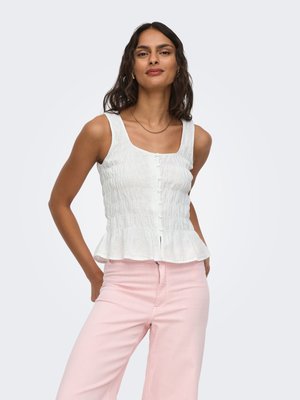 Femme aux longs cheveux foncés portant un haut blanc sans manches boutonné et un pantalon rose clair, posant avec les mains dans le dos.