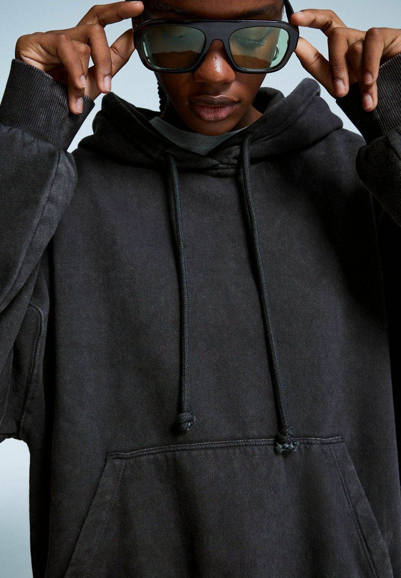 Sort hoodie med snøre, frontlomme og struktureret stof. Store, mørke solbriller i rektangulær form båret i ansigtet.