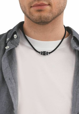 Primo piano del volto inferiore e del collo di un uomo che indossa una collana nera intrecciata con un ciondolo metallico cilindrico sopra una maglietta bianca e una camicia grigia.