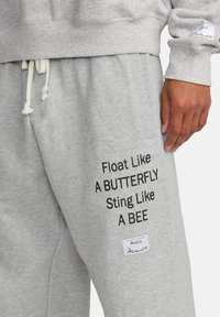 Pantaloni del sudore grigi con coulisse bianche e testo nero che recita "Float Like A BUTTERFLY Sting Like A BEE" vicino alla tasca destra.
