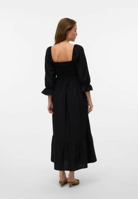 Robe longue noire avec un dos smocké, décolleté carré et manches bouffantes. Comprend un ourlet volanté et un tissu léger.