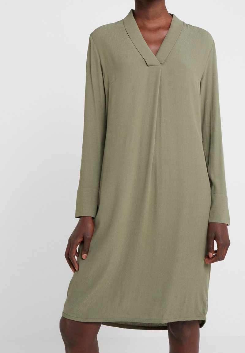 Femme portant une robe olive verte ample, longueur genou, à manches longues et col en V, debout devant un fond clair uni.
