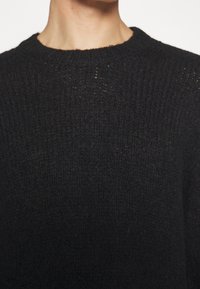 Czarny sweter z dzianiny o wzorze teksturowym, okrągłym dekolcie i luźnym kroju. Materiał wydaje się miękki i ciepły, z widocznymi detalami szycia.