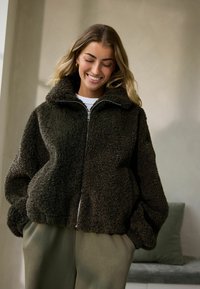 Dunkelbraune, gepolsterte, mit Reißverschluss versehene Jacke mit hohem Kragen und lockerer Passform, kombiniert mit olivgrünen Hosen in einem minimalen Innenraum.