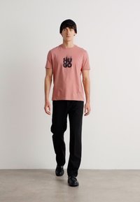 Īsām piedurknēm rozā t-krekls ar melnu "HUGO" logo, apvienots ar melnām biksēm. Modelis ģērbts melnā cepurē un melnās kurpēs, soļojot pret balto fona.