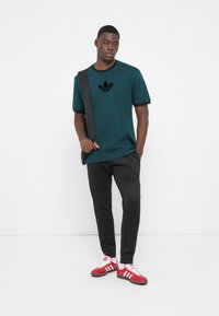 Mørk teal t-shirt med sort kant og et sort logo, parret med sorte bukser og hvid sneakers med røde striber. Modellen bærer en mørk taske.